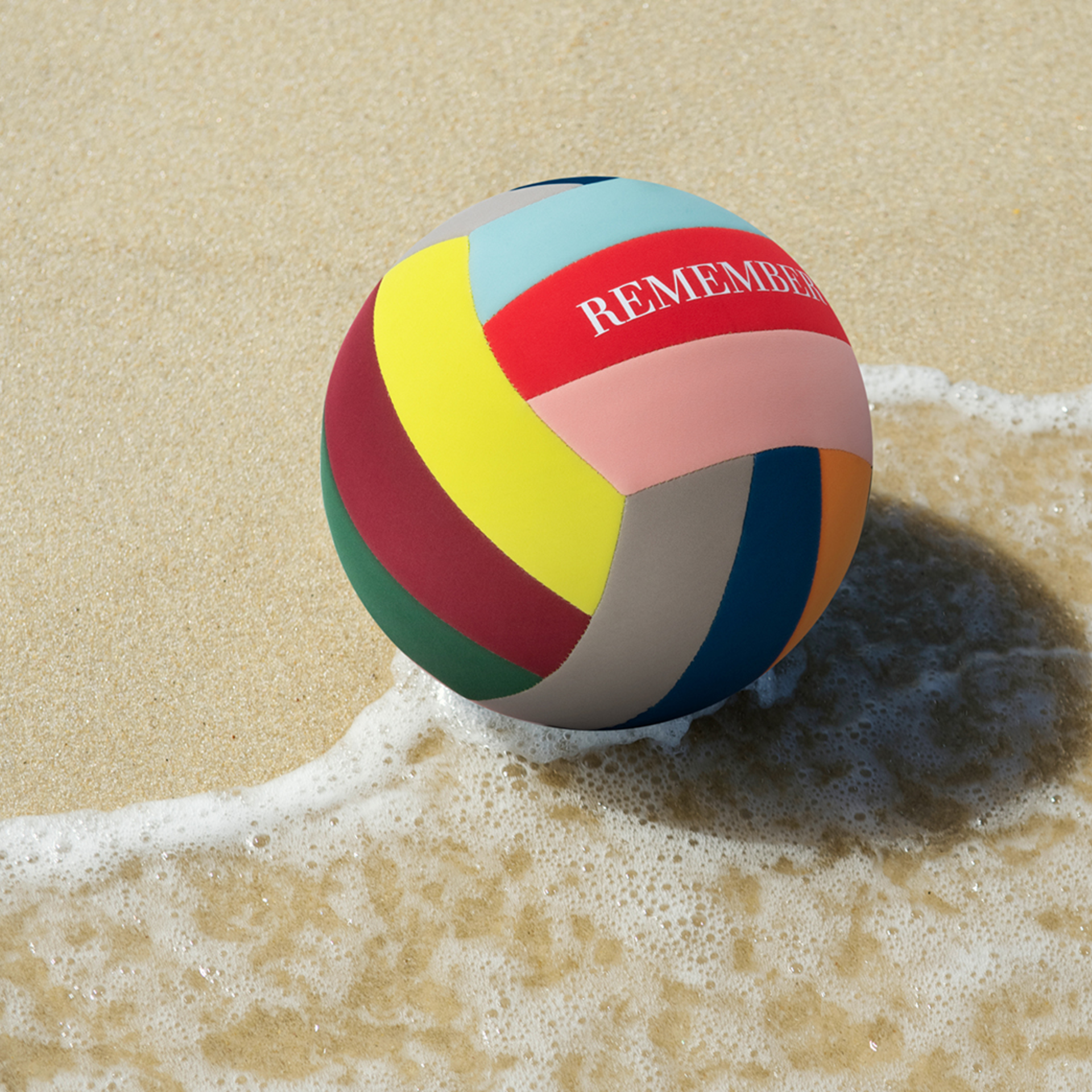 Ein bunter Freizeit-Ball liegt am Strand, teilweise im hellen Wasser. Auf einem roten Segment steht der Schriftzug "REMEMBER".
