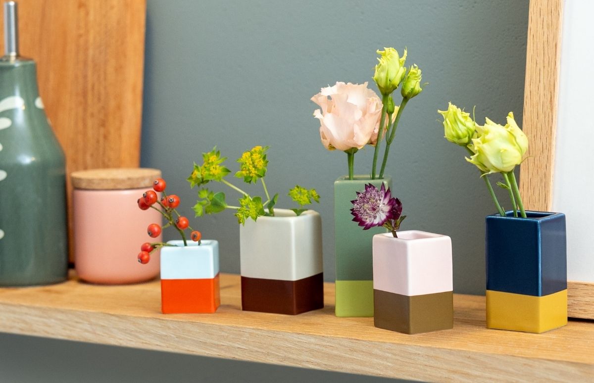 Fünf zweifarbige Keramikvasen auf einem Holzregal. Sie enthalten: rote Beeren in orange-hellblauer Vase, gelbgrüne Blüten in braun-cremefarbener, rosa Rose in grüner, lila Blume in pink-olivgrüner. Graue Wand.