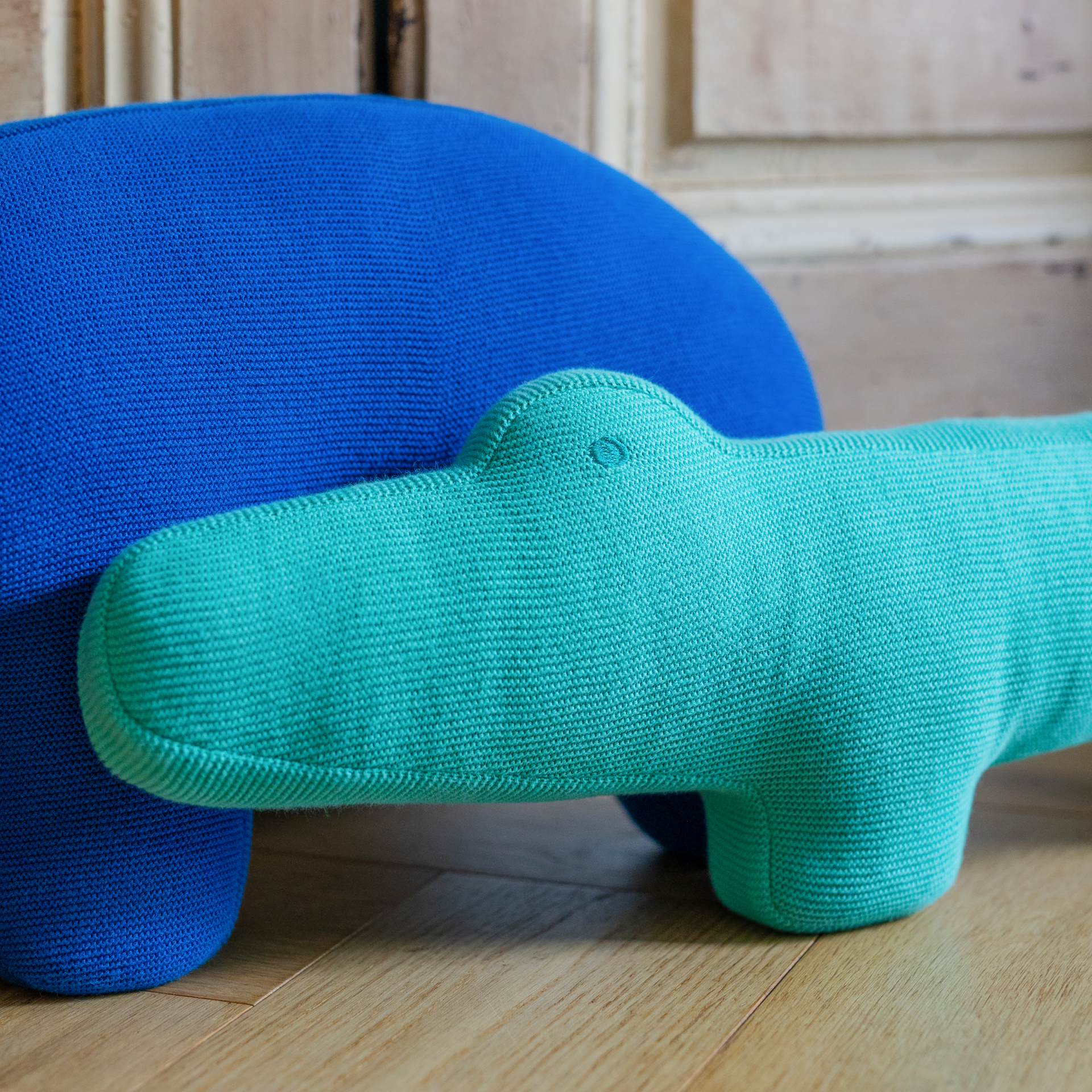 Ein türkis-farbenes Kissen in Krokodilform, „Kroko Karl“, liegt vor einem blauen, runden Kissen auf hellem Holzboden.
