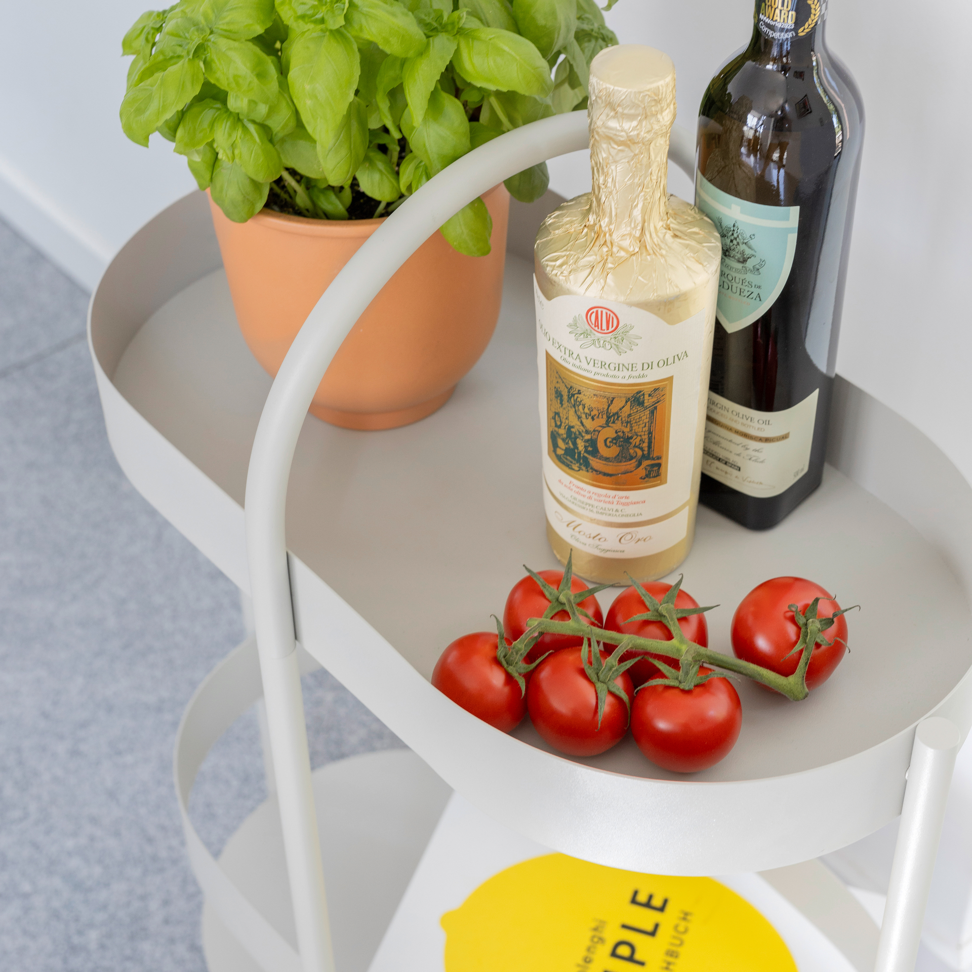 Helle Beistellkonsole Barbara mit Basilikum, Olivenöl, Tomaten und einem Buch. Zwei Ebenen und ein Griff sind erkennbar.