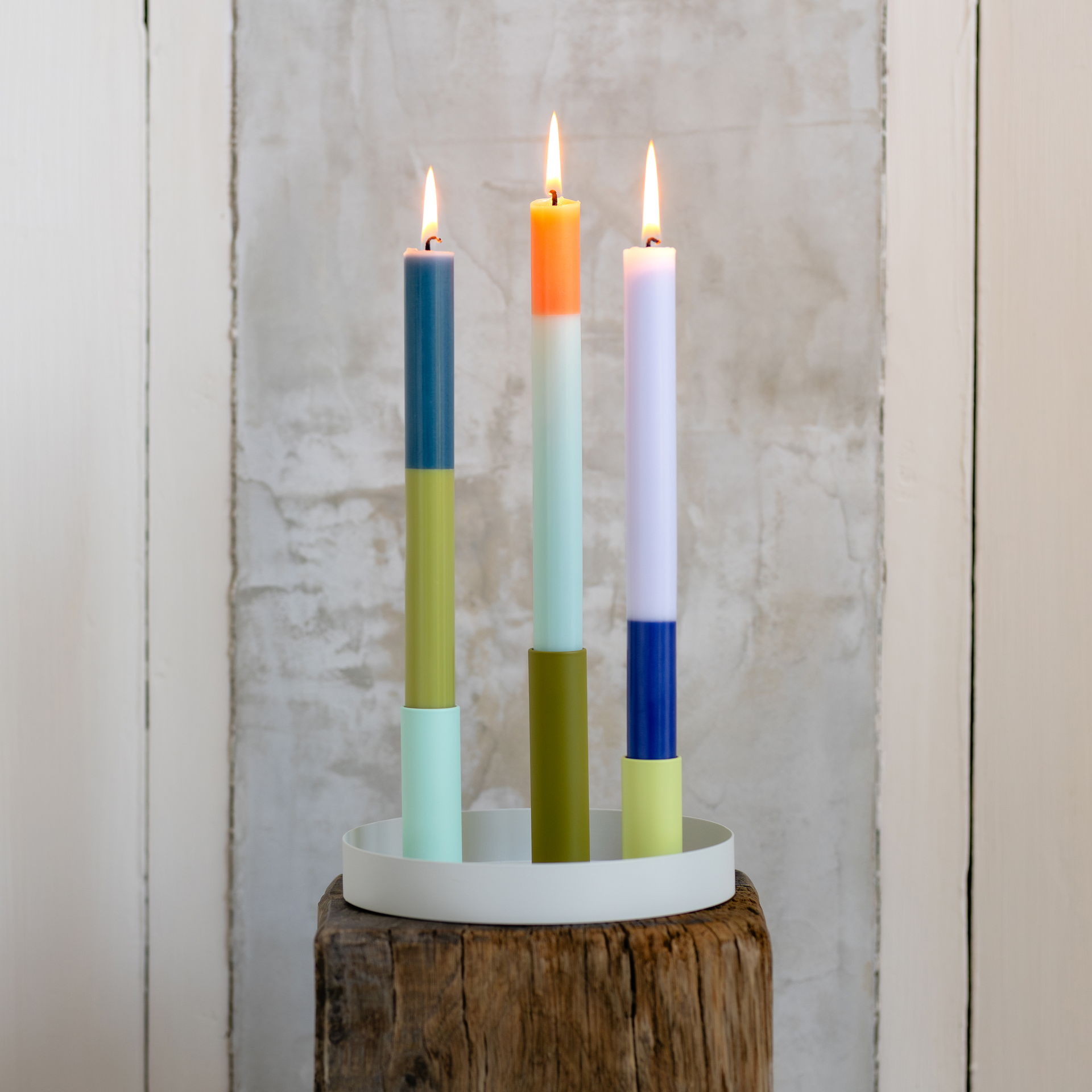 Drei bunte Stabkerzen, in magnetischen Kerzenhaltern, stehen auf einem weißen Tablett. Die Kerzen sind angezündet. Das 'Luna Mini' Kerzenhalter Set steht auf einem Holzschemel.