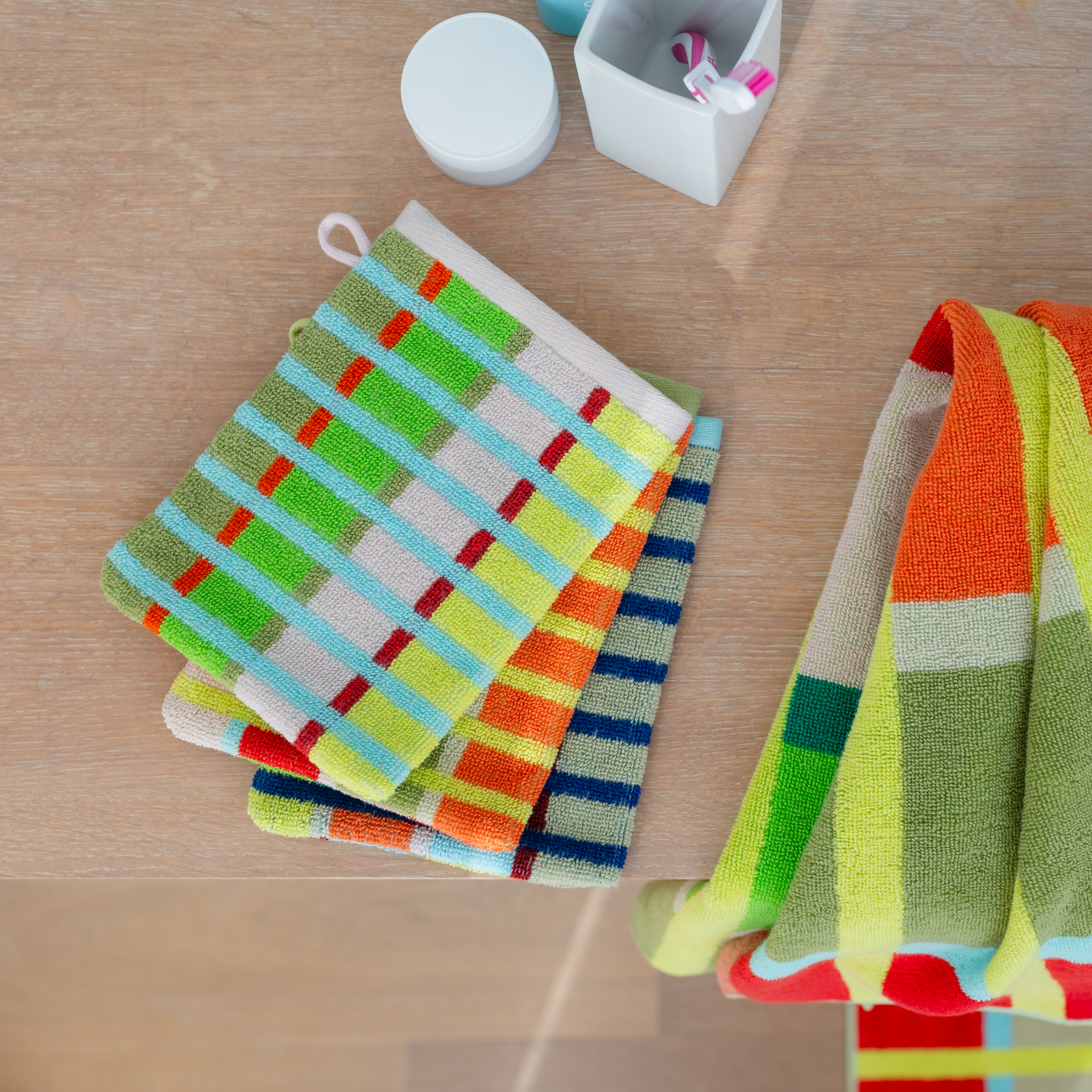 Trois gants de toilette Kaido disposés côte à côte sur un fond blanc, avec un motif à rayures et une boucle de suspension : à gauche vert-jaune-rose-rouge, au centre bleu-jaune-marron, à droite des tons pastel : bleu clair, rose, rouge, vert.