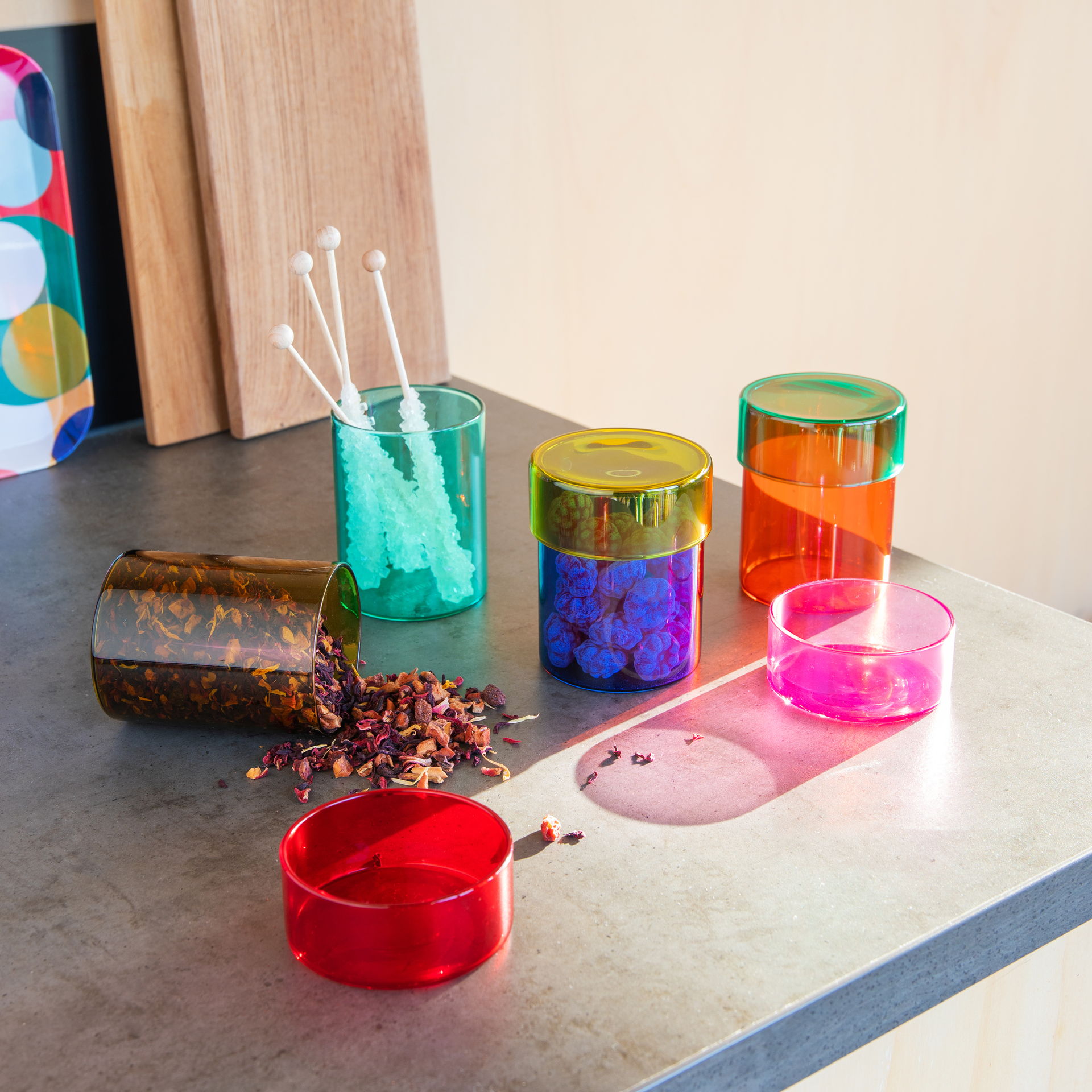 Glasdosen-Set 'Cosima' in verschiedenen Farben und mit diversen Inhalten auf einer grauen Oberfläche, ein Behälter ist umgefallen und der Inhalt liegt daneben.