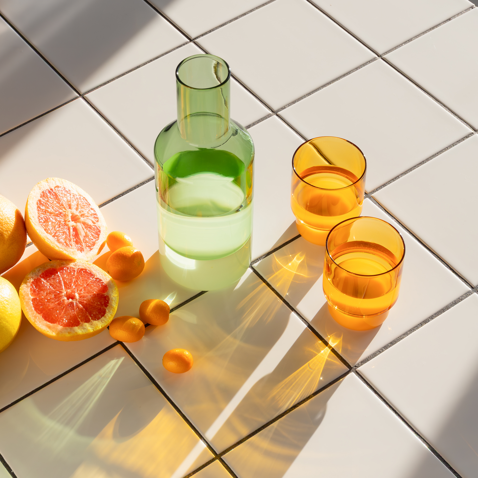 Auf weißen Fliesen stehen eine grüne Karaffe, zwei orangefarbene Gläser, halbe Grapefruits und Kumquats im hellen Sonnenlicht.