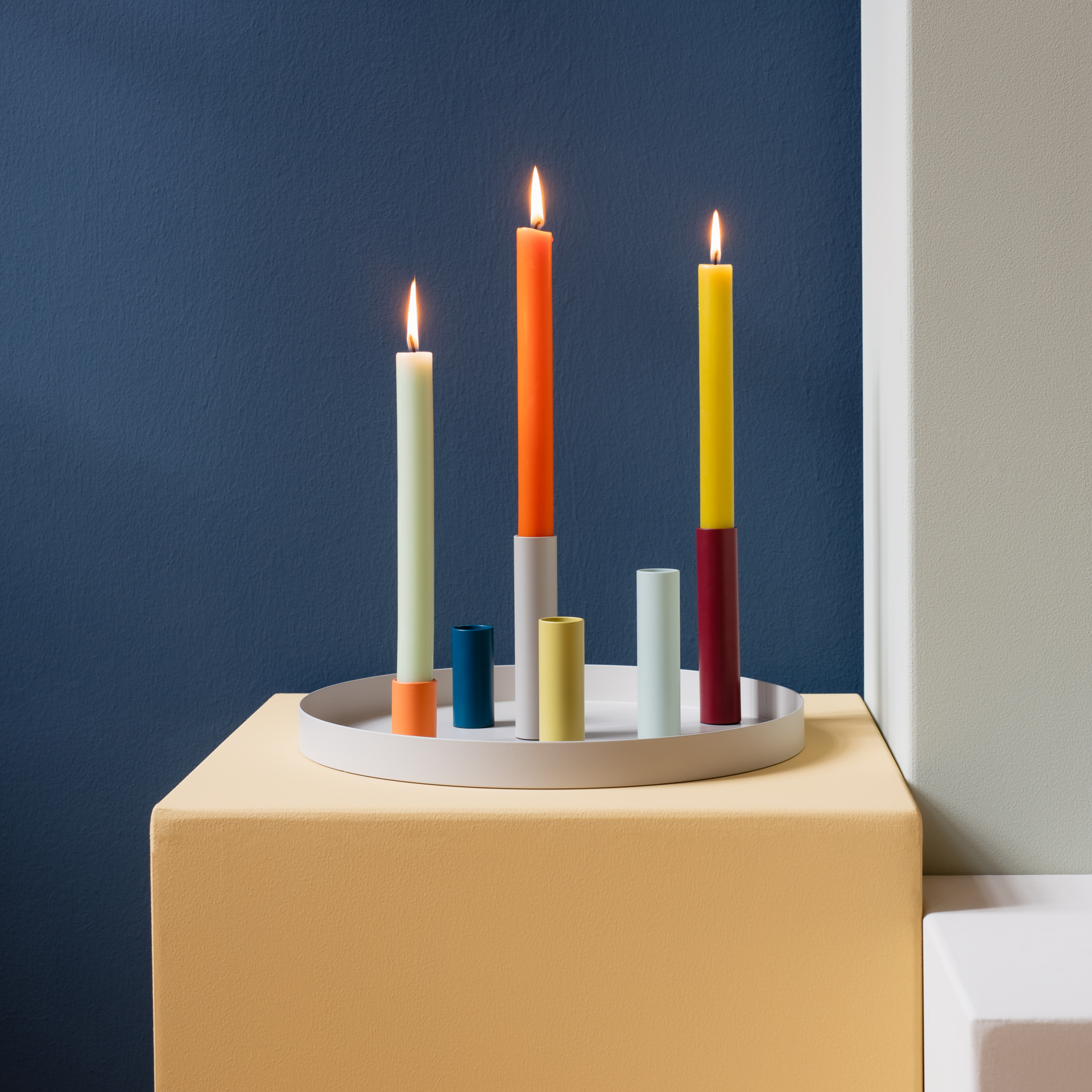 Ein Arrangement des magnetischen Kerzenhalters "Luna" mit farbigen Kerzen auf einem Tablett vor einer blauen Wand. Drei Kerzen brennen.
