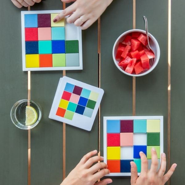 Deux personnes font des puzzles de couleurs sur une table en bois vert foncé avec bandes cuivrées. Une main place une pièce jaune clair en haut, deux mains s'activent en bas. À côté : pastèque, eau citronnée, nuancier.