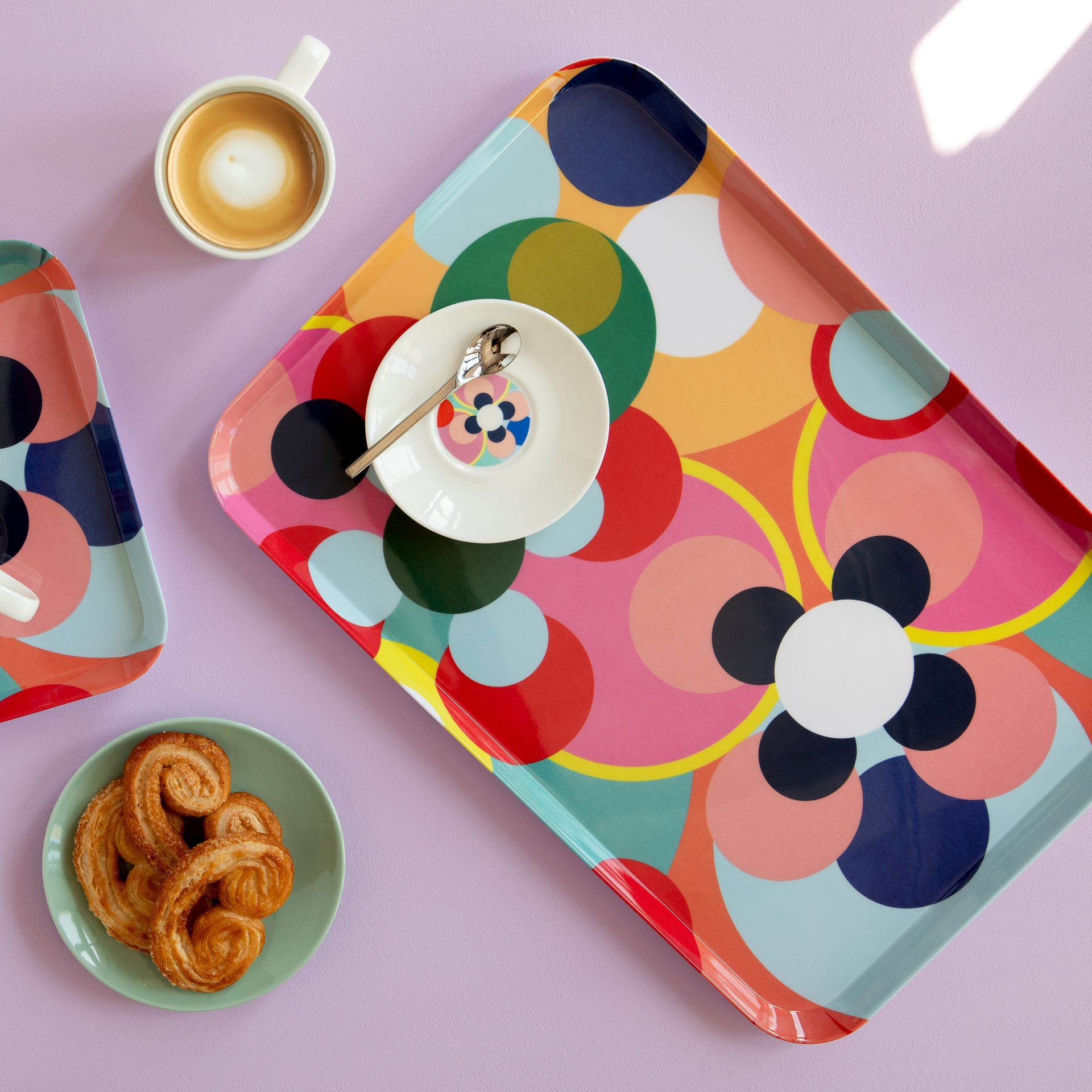 Ein farbenfrohes 'Sushi' Tablett mit Cappuccino, Gebäck und Untertasse auf lila Hintergrund. Das Tablettmuster besteht aus bunten Kreisen und Blütenformen.