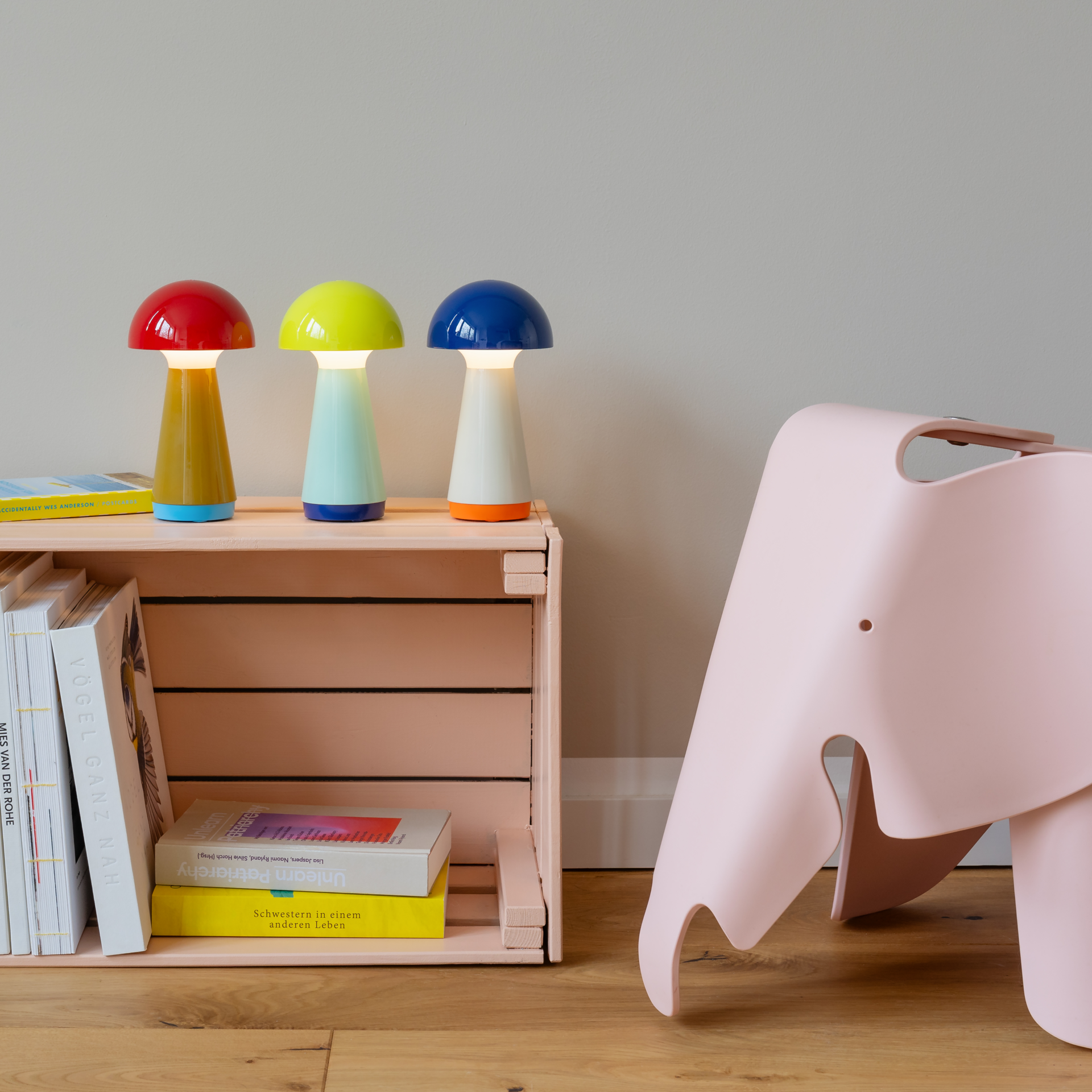 Drei 'Bobbi' Tischleuchten in blau, rot und gelb auf einem Regal mit Büchern, daneben ein rosa Elefanten-Hocker auf einem Holzboden.