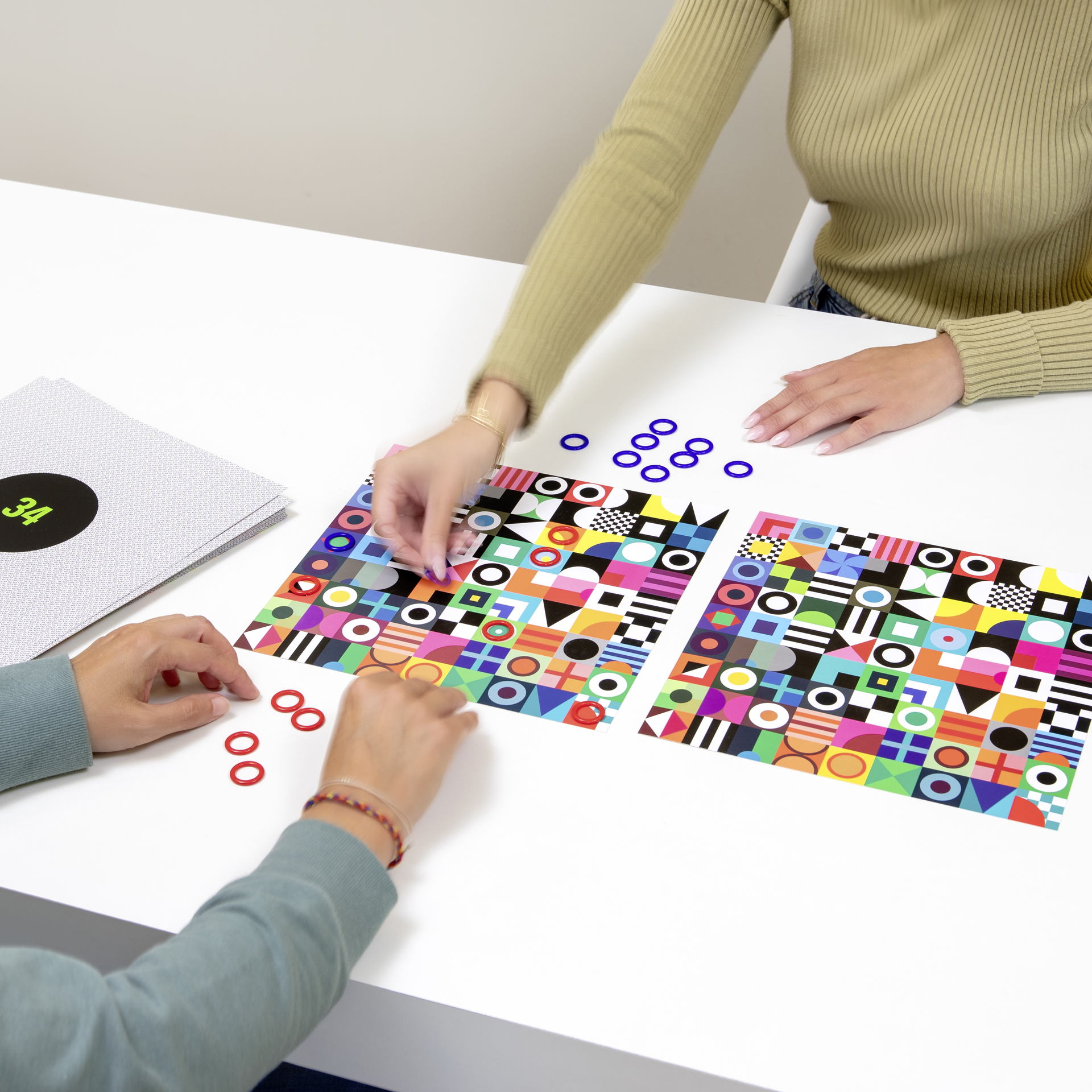 Zwei Personen spielen das „Double“-Suchspiel. Bunte Spielkarten mit geometrischen Mustern, rote und blaue Ringe liegen bereit.