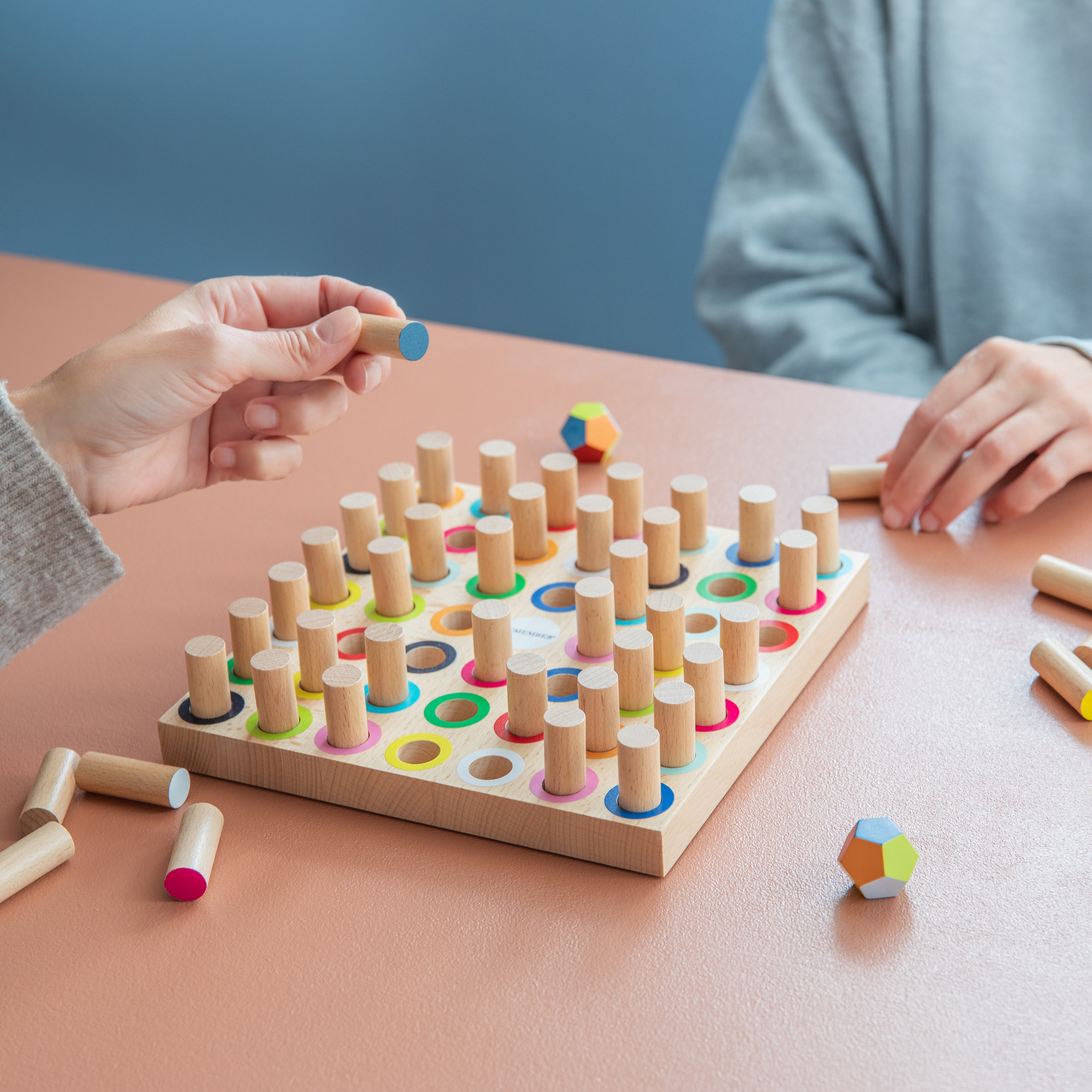 Holzspiel Momo: Holzbrett mit farbigen Kreisen und Holzstiften. Ein farbiger Polyeder und weitere Stifte liegen daneben. Oben links steht "MOMO" in grossen Buchstaben.
