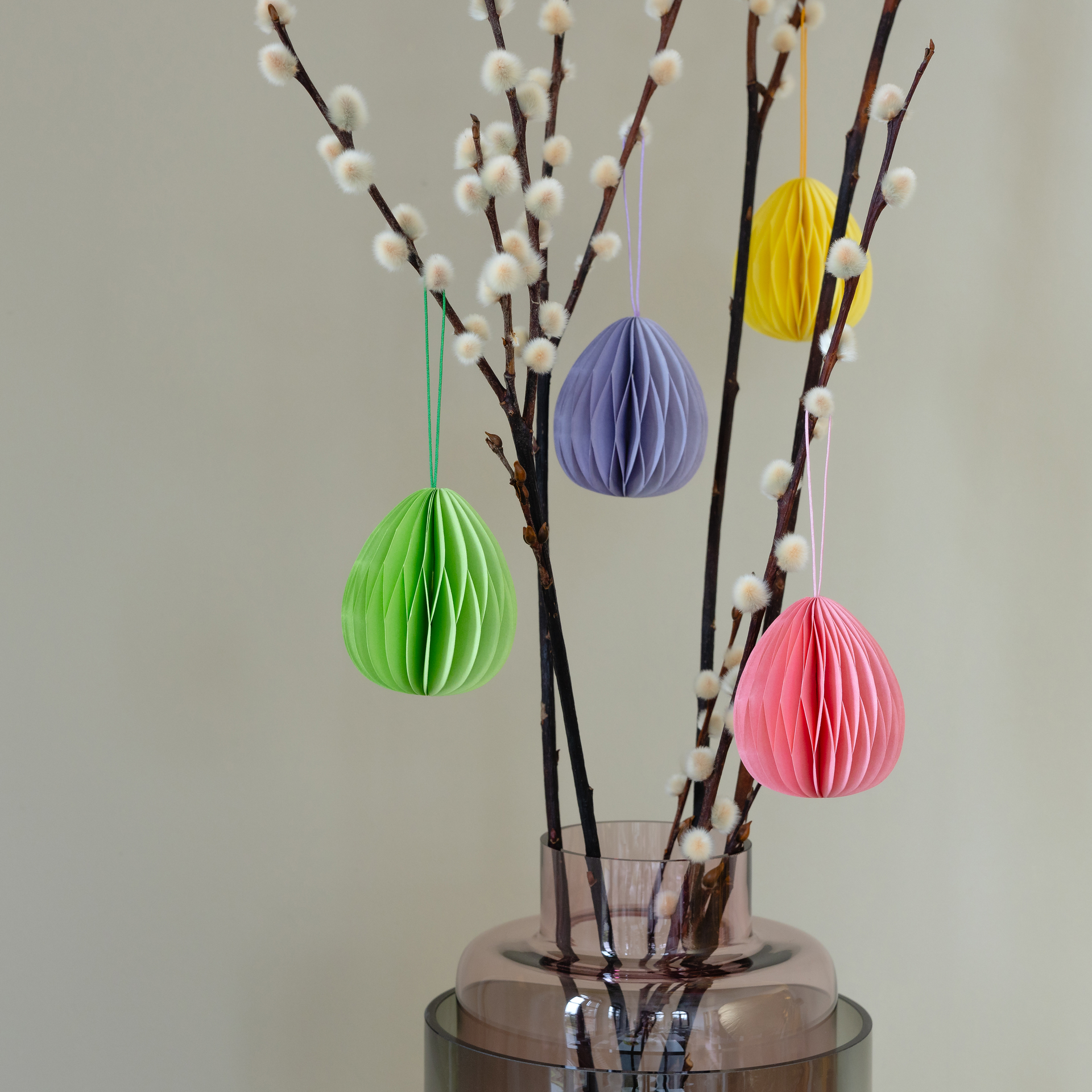 Zarte Weidenkätzchen in Vase mit vier farbigen, ovalen Baumschmuck-Anhängern aus Papier. Die Farben sind Grün, Lila, Gelb und Rosa.