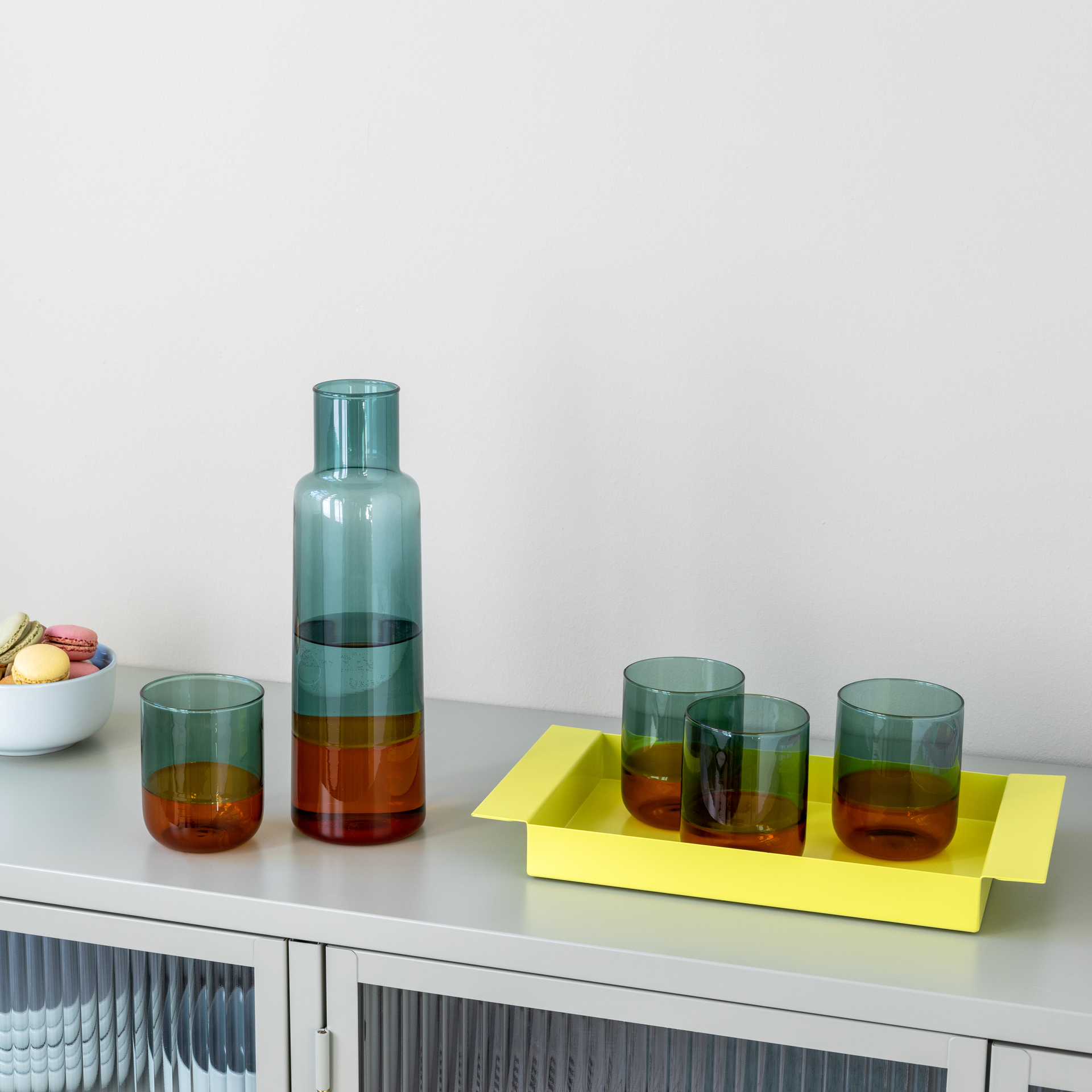 Zwei Gläser 'amber' und eine Karaffe auf einer Ablage. Die Trinkgläser sind zweifarbig: Grün und braun. Ein gelbes Tablett und eine Schale mit Macarons sind auch zu sehen.