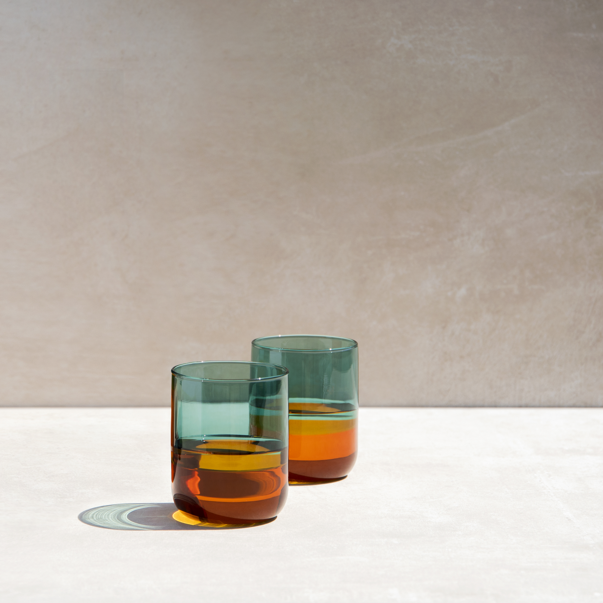 Zwei hohe, schlanke Gläser im Amber-Design stehen auf einer hellen Fläche vor einem beigen Hintergrund. Die Gläser sind teilweise mit einer bernsteinfarbenen Flüssigkeit gefüllt.