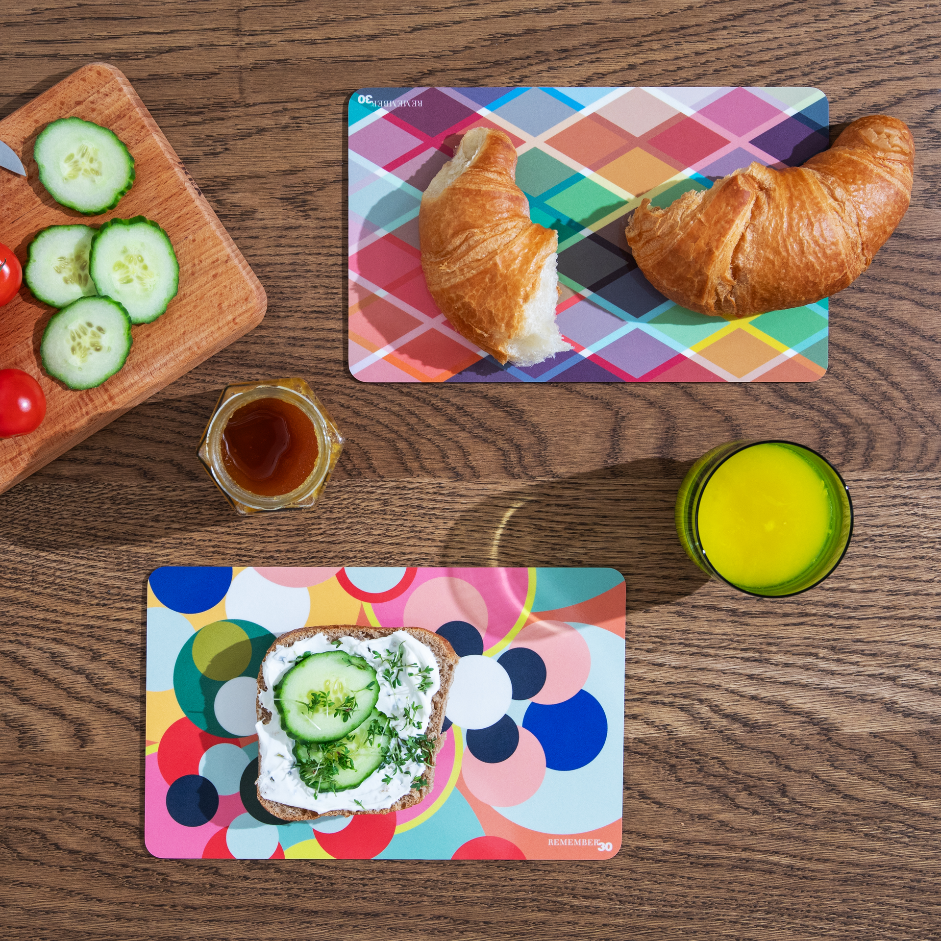Colazione su un tavolo di legno: pane con cetrioli, croissant, miele, succo di frutta, fette di cetriolo e di pomodoro su una tavola. Sottofondi colorati tra i piatti.