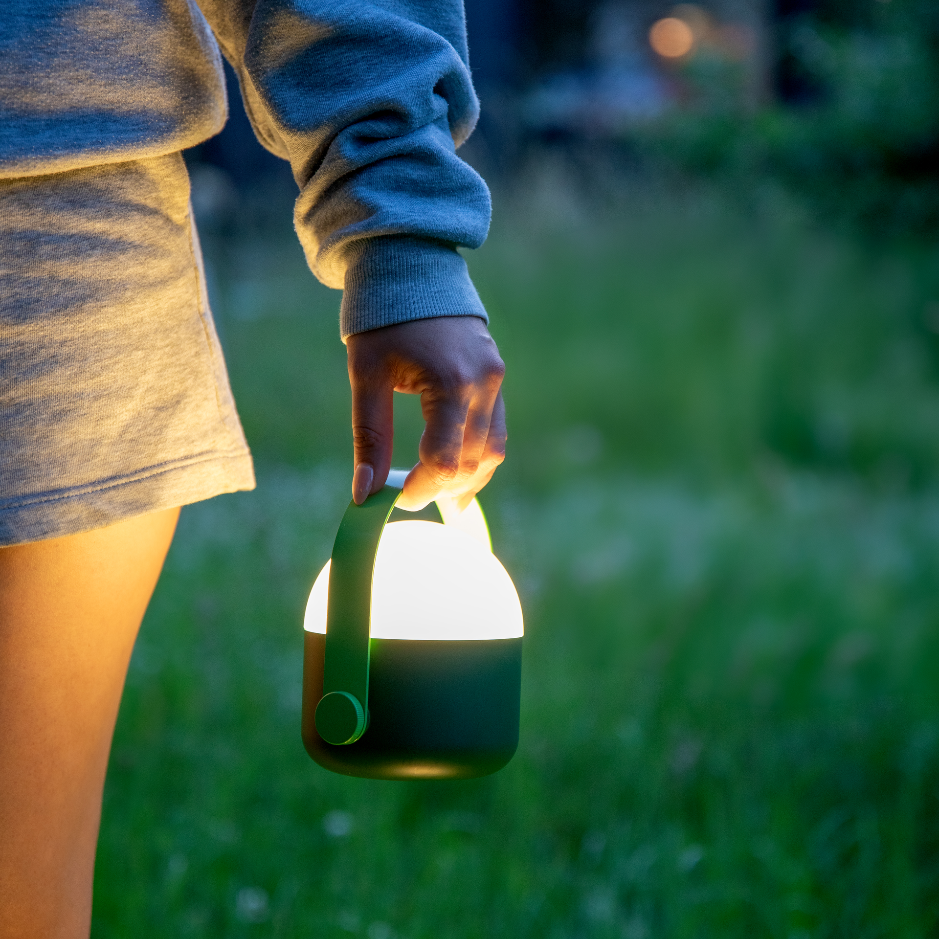 Eine Person hält eine leuchtende, tragbare grüne Laterne in der Hand. Der Hintergrund ist ein verschwommener grüner Garten bei Dämmerung.