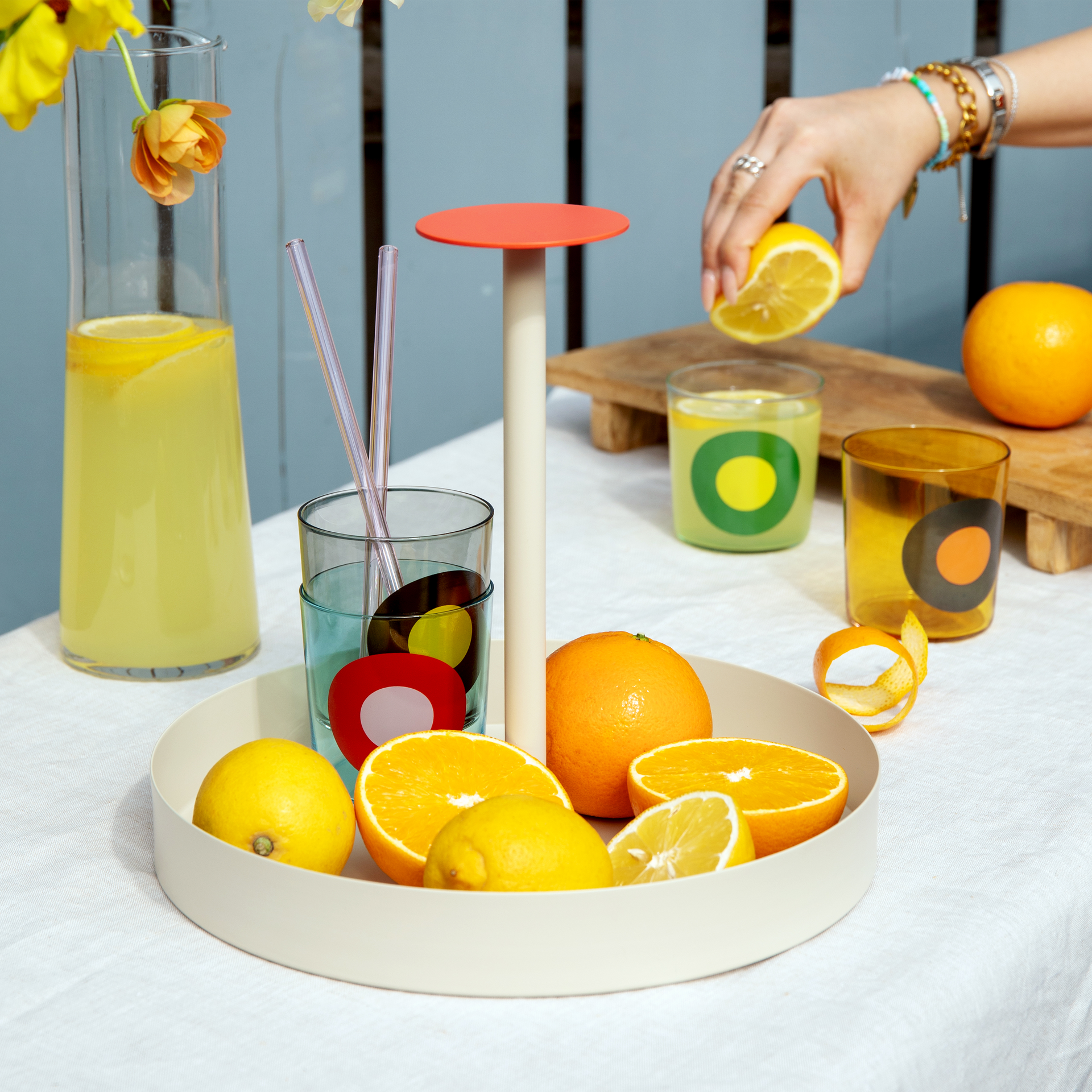 Nature morte avec des agrumes et des boissons sur une table blanche. Une main presse un citron au-dessus d'un verre. Un bouquet de fleurs et deux verres décoratifs sont visibles.
