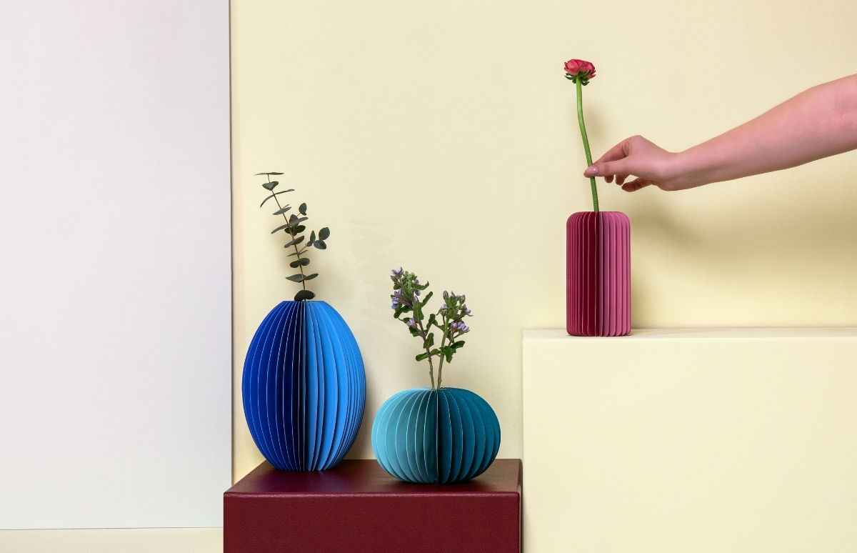 Plissierte Papiervasen mit Pflanzen: große blaue ovale Vase mit Eukalyptus, kleine türkisfarbene runde Vase mit violetten Blüten und pinkfarbene rechteckige Vase, in die eine Hand eine rote Ranunkel stellt.