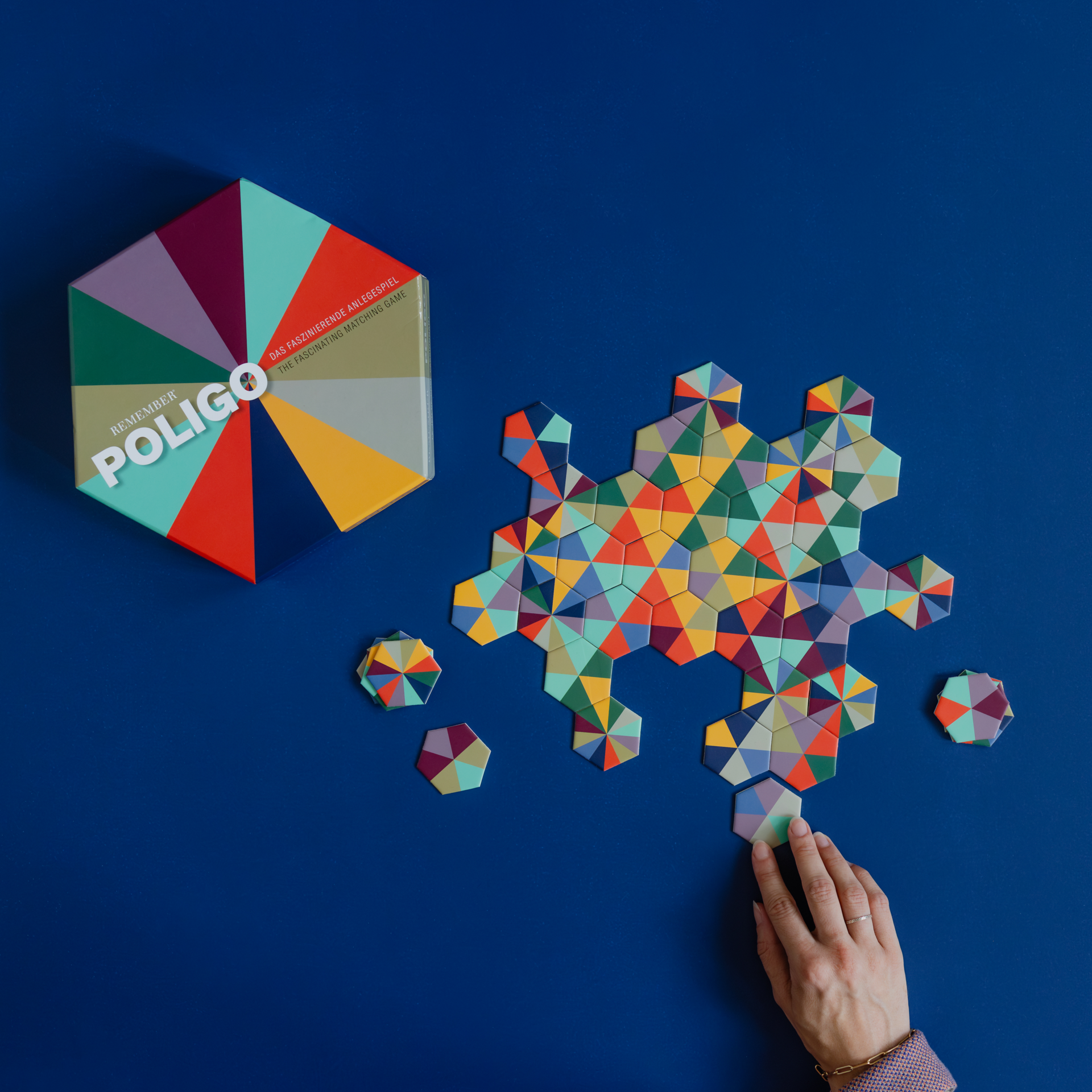 Draufsicht auf das Poligo Spiel mit Spielkarten und einer Hand auf blauem Grund. Farbiges, sechseckiges Spiel-Puzzles.
