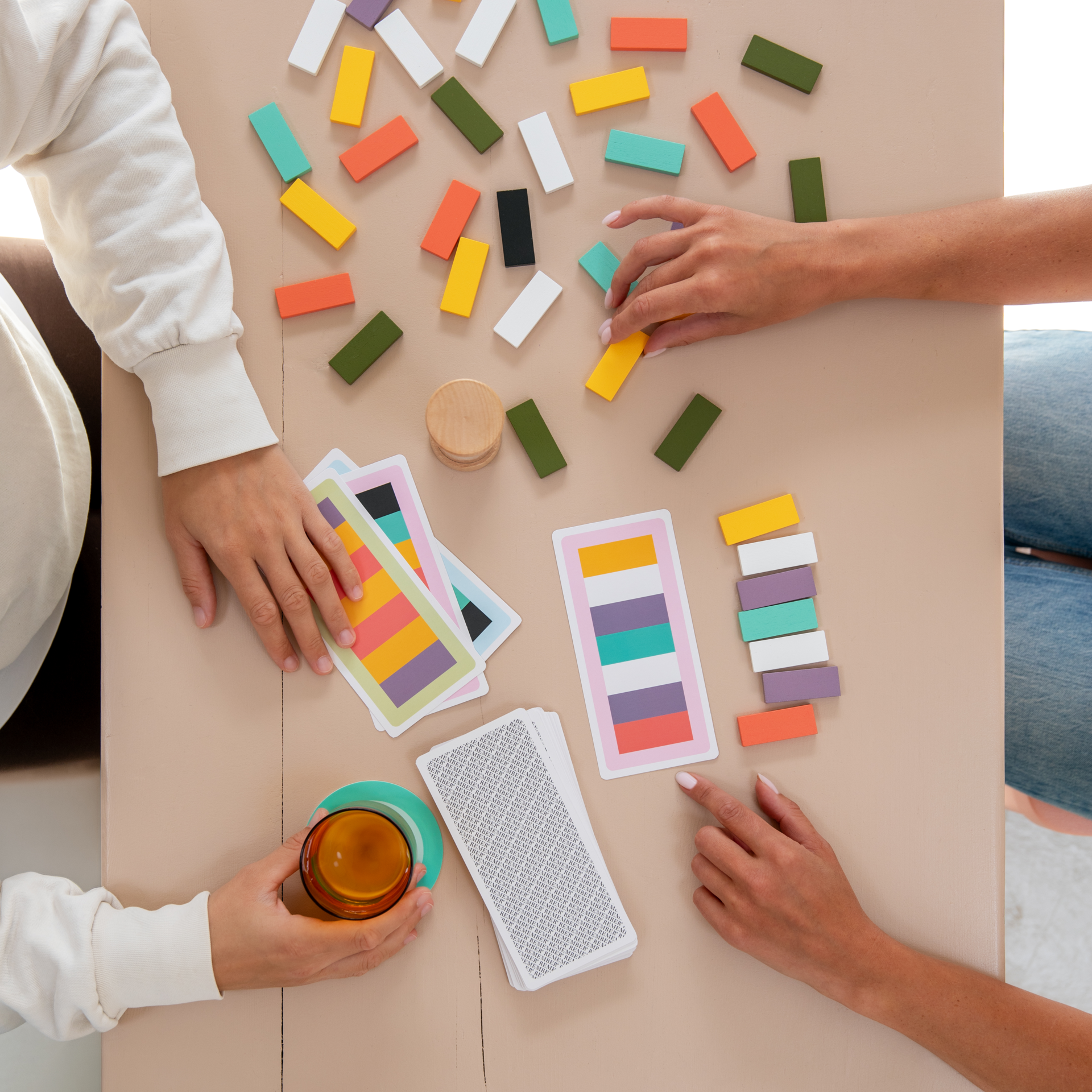 Deux joueurs jouent au jeu de mémoire Rigato autour d'une table, avec des tuiles en bois en sept couleurs. L'un tient des cartes mission à motifs colorés, l'autre pose les tuiles. Une pile « REMEMBER » est face cachée.