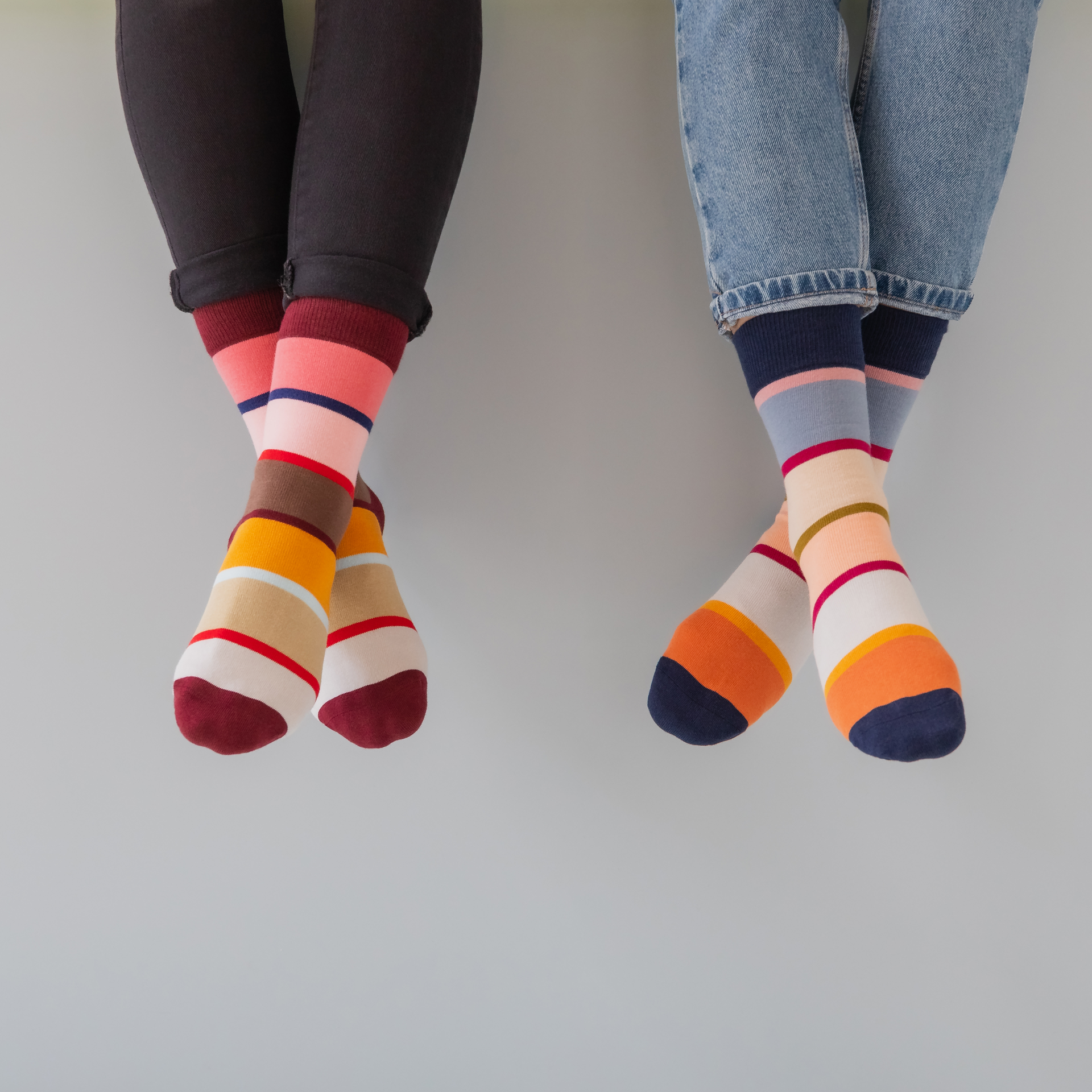 Zwei gekreuzte Beinpaare mit farbenfrohen, gestreiften Socken. Ein Bein in schwarzer Hose, das andere in Jeans. Die Socken sind geringelt in diversen Farben.