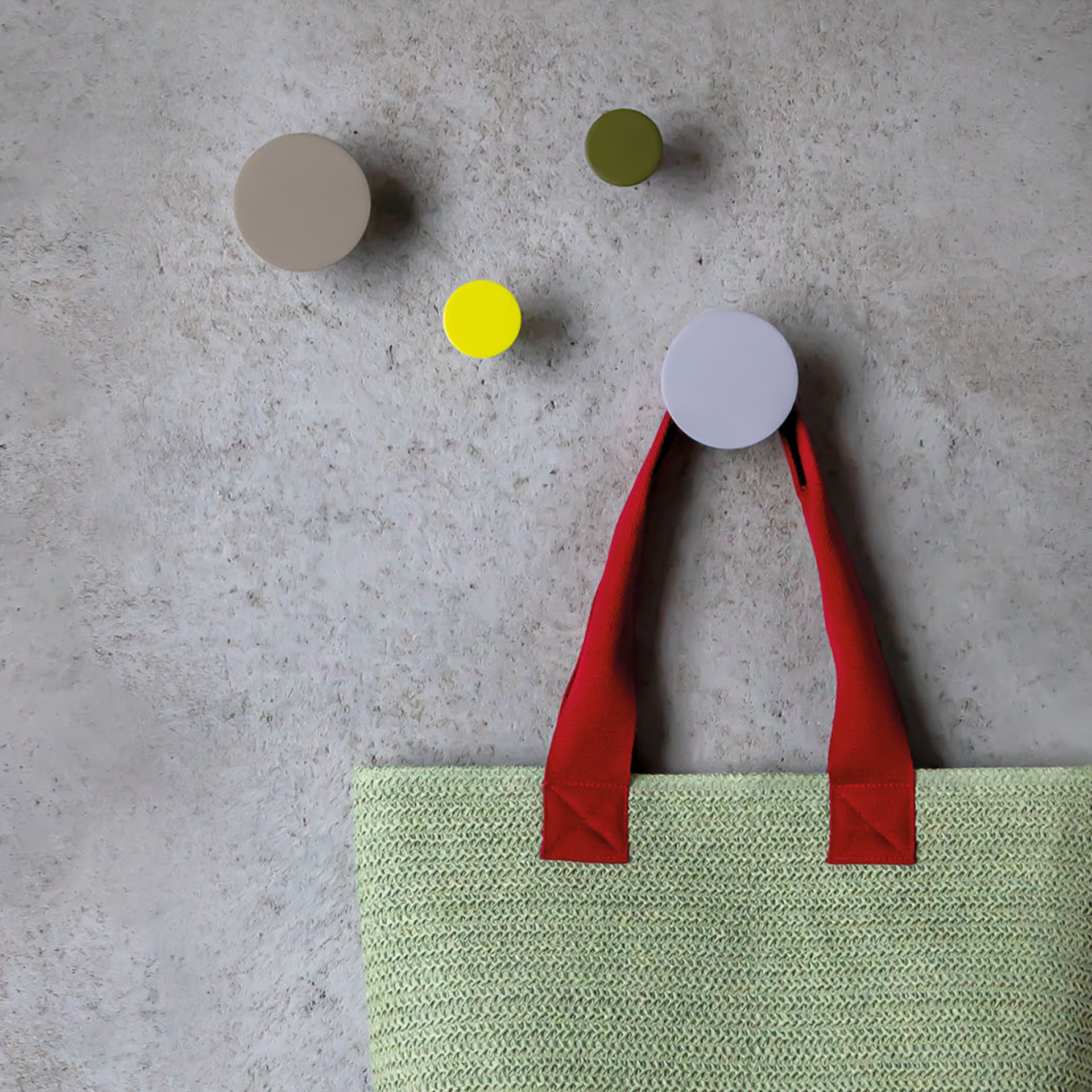 Vier Wandhaken in Beige, Grün, Gelb und Lila an einer Wand, wobei an einem Haken eine hellgrüne Tasche mit roten Henkeln hängt.