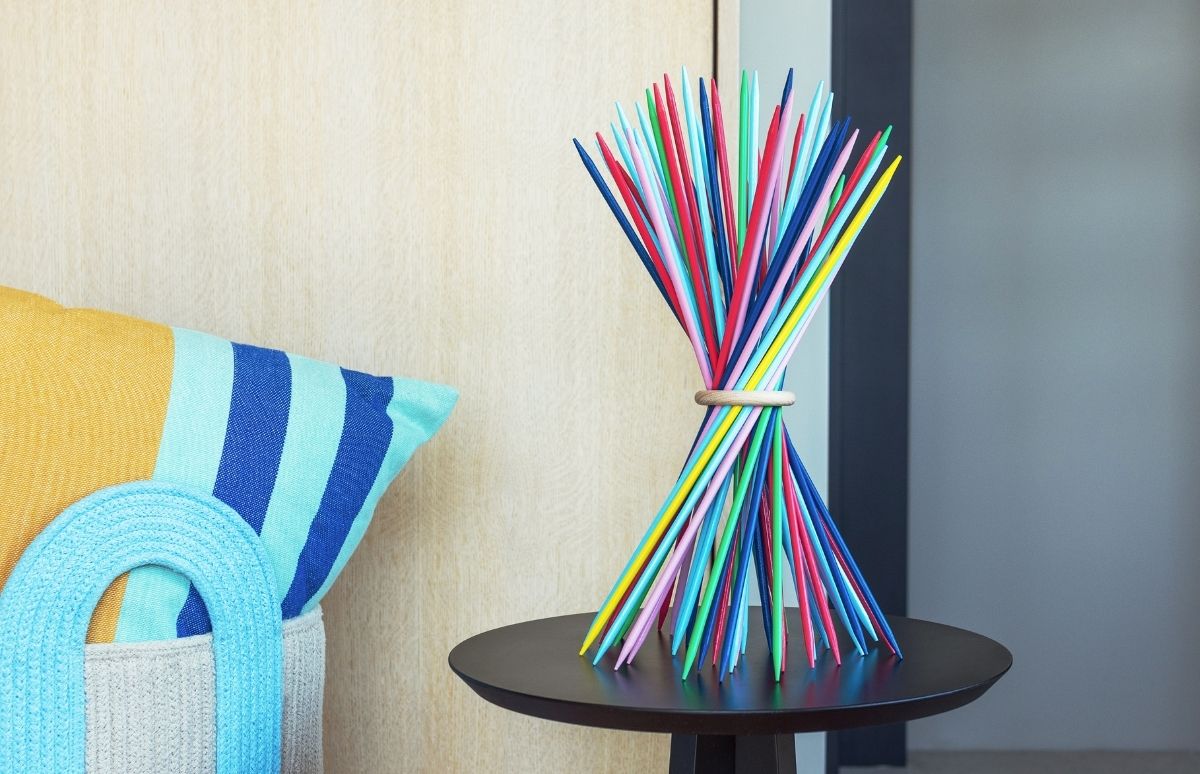 Un bouquet coloré de bâtonnets de Mikado, attachés par un anneau en bois, s'épanouit sur une table d'appoint foncée. À gauche, un coussin rayé jaune/bleu, puis un sac clair à anse bleue. Mur bois clair et gris foncé.