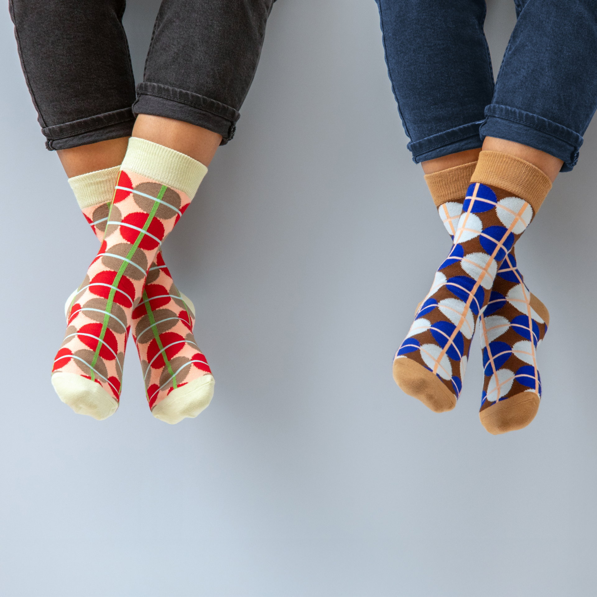 Zwei gekreuzte Beinpaare mit Socken in Muster und Farbe, einmal rot/braun/grün und einmal blau/braun/weiß, jeweils passend zur Jeansfarbe.