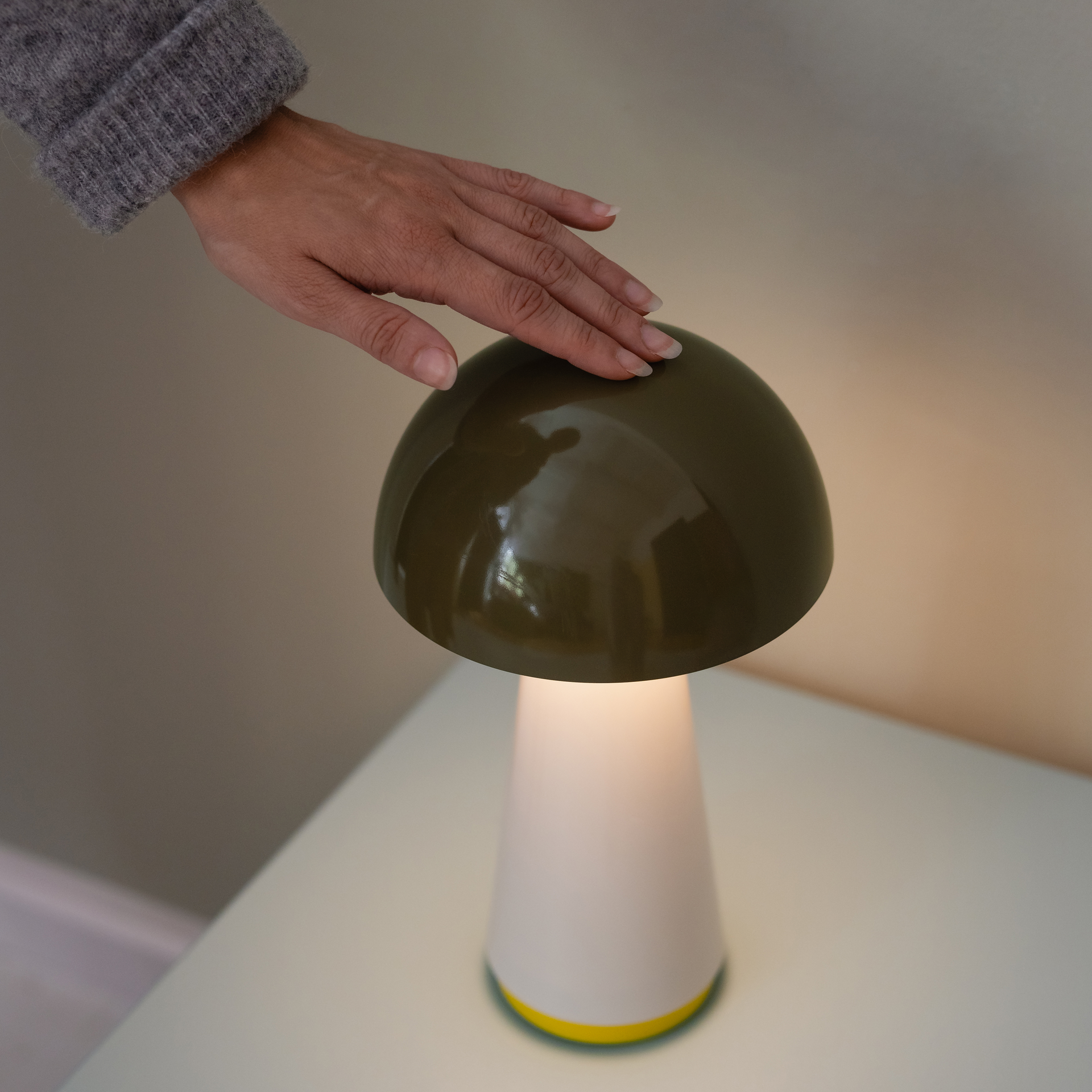 Nahaufnahme der olivgrünen Tischleuchte Bob. Eine Hand berührt den Lampenschirm. Das Licht der Leuchte ist warmweiß und scheint dezent.