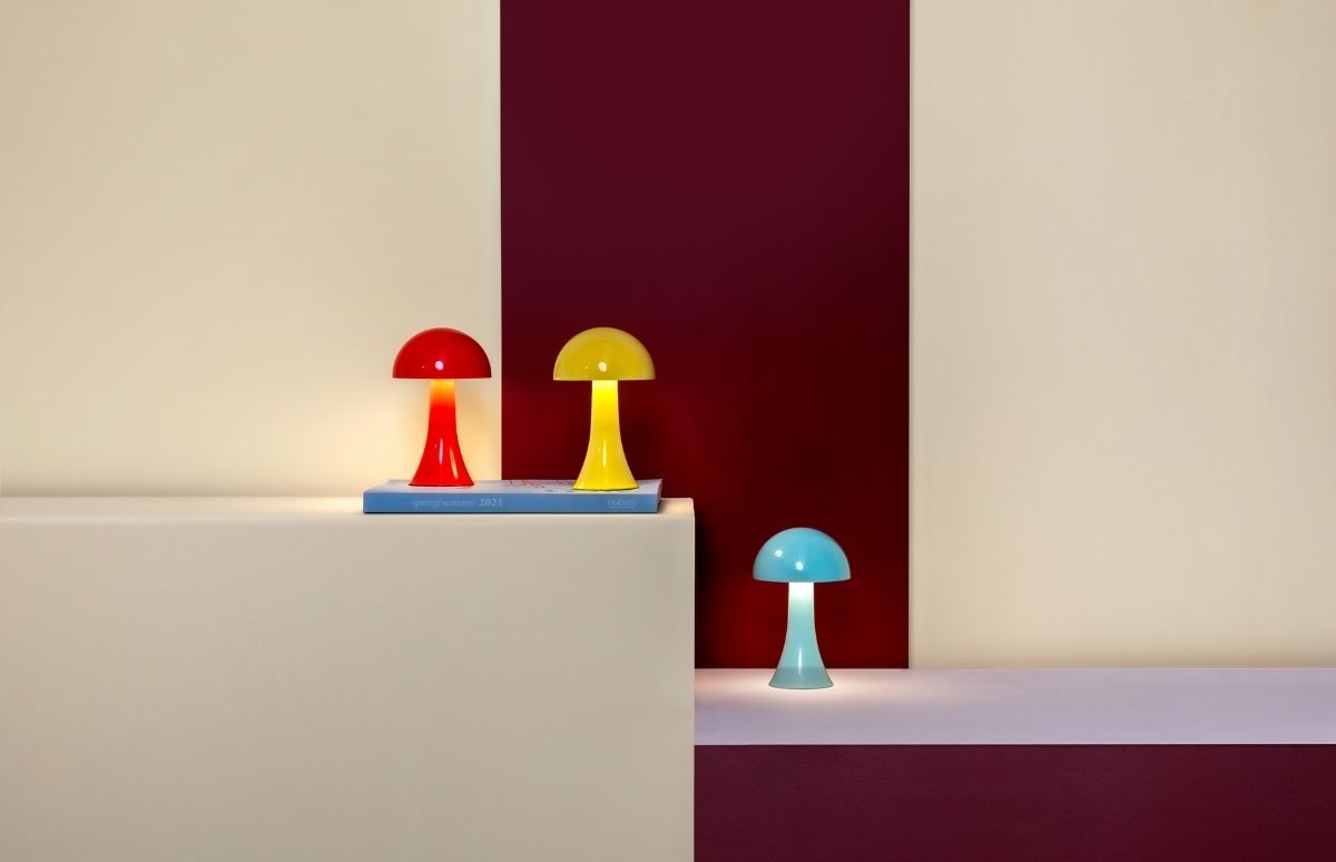 Drei leuchtende Pilzlampen in Rot, Gelb und Hellblau stehen auf unterschiedlich hohen, farblich abgesetzten Flächen. Rot und Gelb auf Beige über einem blauen Buch, Hellblau auf Lila. Hintergrund: Creme und bordeauxroter Streifen.