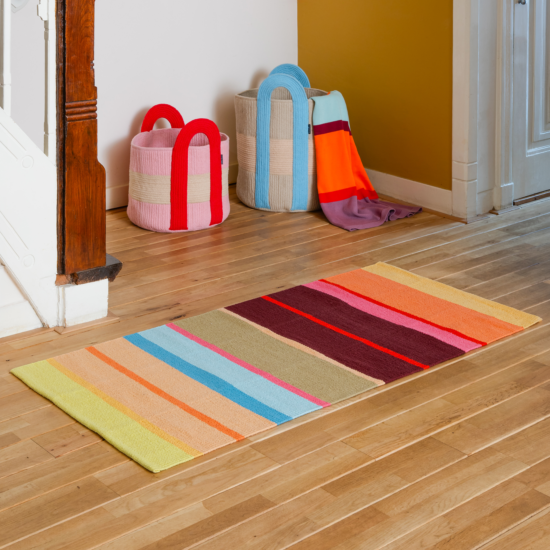 Tapis de couloir coloré à rayures horizontales jaunes, orange, rose, bleu clair, vert olive et bordeaux, posé sur un parquet clair. Texture du coton tissé à la main bien visible. À l'arrière-plan, des paniers et une couverture pliée.